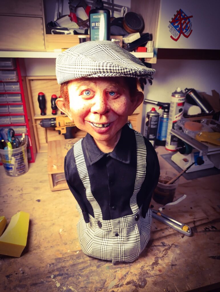 The completed Alfred E. Neuman bust by sculptor Pierre Schrader, wearing a matching houndstooth flat cap, shirt, and suspenders in his workshop, with the traditional stonemason guild symbol visible on the cabinet above.