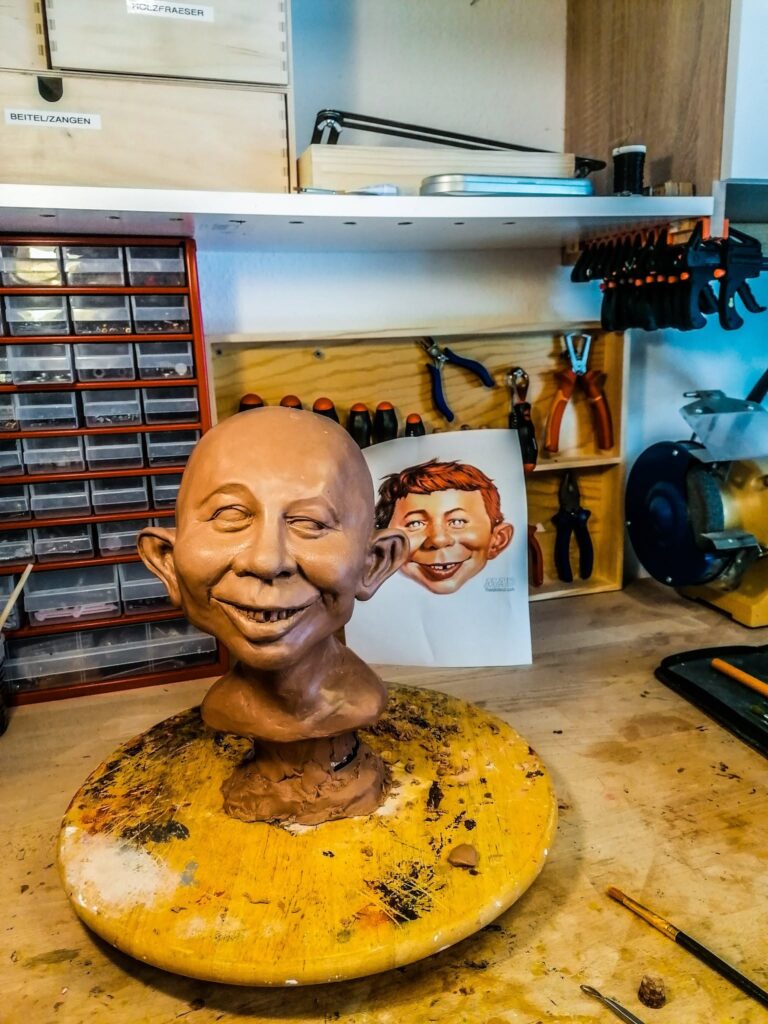 A raw brown clay sculpture of an Alfred E. Neuman bust on a yellow turntable in a workshop, with Jordu Schell’s reference printout pinned to the wall in the background.