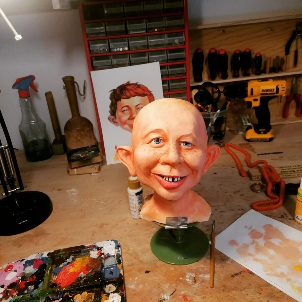 The unhaired cast of an Alfred E. Neuman bust being hand-painted on a workbench, featuring realistic skin tones, freckles, bright blue eyes, and the signature gap-toothed smile.