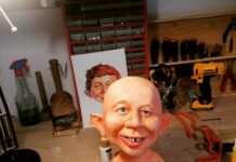 The unhaired cast of an Alfred E. Neuman bust being hand-painted on a workbench, featuring realistic skin tones, freckles, bright blue eyes, and the signature gap-toothed smile.