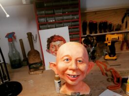 The unhaired cast of an Alfred E. Neuman bust being hand-painted on a workbench, featuring realistic skin tones, freckles, bright blue eyes, and the signature gap-toothed smile.