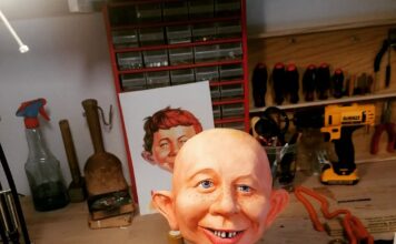 The unhaired cast of an Alfred E. Neuman bust being hand-painted on a workbench, featuring realistic skin tones, freckles, bright blue eyes, and the signature gap-toothed smile.