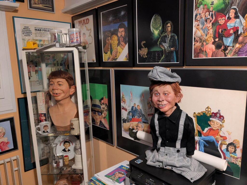 The handmade Alfred E. Neuman bust by Pierre Schrader in a houndstooth outfit placed next to a glass display case containing the rare Warner Bros. bust by Gary Mirabelle, surrounded by framed MAD Magazine artwork.
