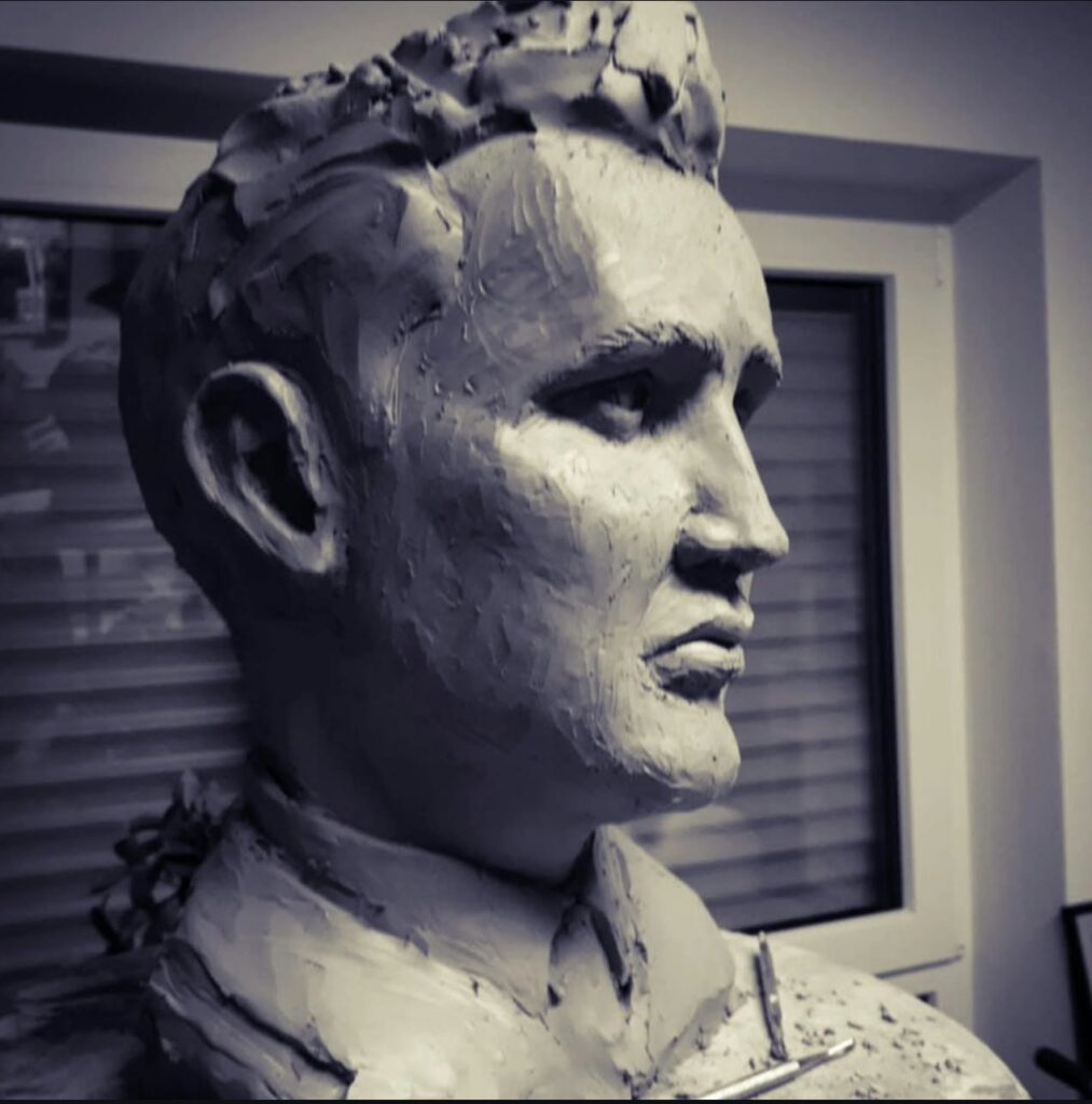 A black and white photograph of an in-progress portrait bust of Elvis Presley sculpted in raw clay, showing rough textures and modeling tools.