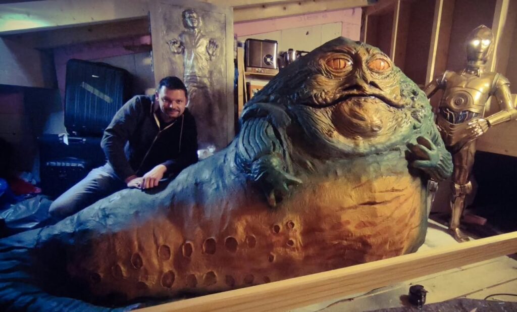 German sculptor Pierre Schrader posing in his workshop next to a massive, life-size sculpture of Jabba the Hutt, flanked by an illuminated C-3PO and a Han Solo in Carbonite wall panel.